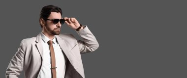 Gri arka planda açık renk takım elbise ve güneş gözlüğü takan yakışıklı sakallı adam.