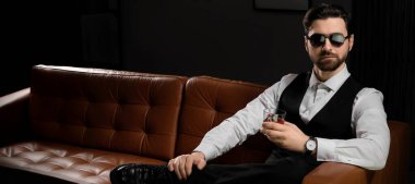 Resmi giyinmiş yakışıklı adam karanlık odada kanepede otururken viski içiyor.