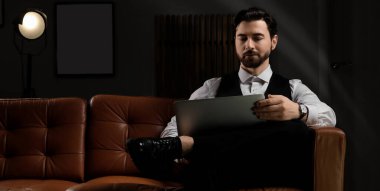 Resmi giyinmiş yakışıklı bir adam ve karanlık odadaki kanepede oturan dizüstü bilgisayarlı.