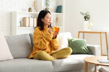 Oturma odasındaki kanepede çay içen güzel, genç Asyalı kadın.