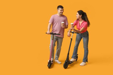 Modern elektrikli scooterları ve sarı arka planda kağıt kahve fincanları olan genç ve güzel bir çift.