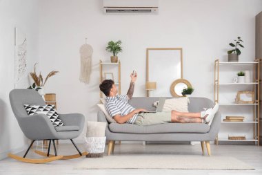 Dizüstü bilgisayarı olan yakışıklı genç adam oturma odasındaki kanepede klimayı çeviriyor.
