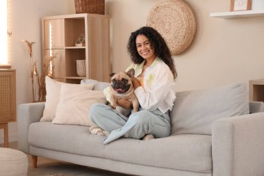 Oturma odasındaki kanepede oturan sevimli Fransız bulldoguyla mutlu genç Afro-Amerikalı kadın.