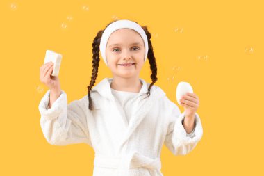 Sabun barları ve sarı arka planda kabarcıkları olan bornozlu küçük bir kız.