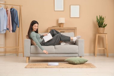 Evde elektrikli süpürgeyle kanepede kitap okuyan genç bir kadın.