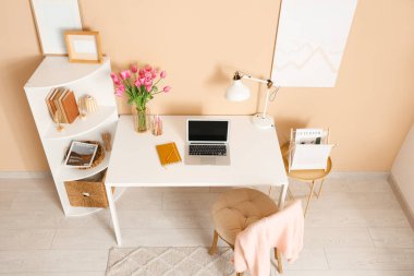 Modern dizüstü bilgisayarı, raf ünitesi ve bej duvarın yanında güzel lale çiçekleri olan lüks çalışma alanı.