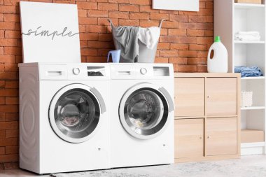 Çamaşır odasının içinde, modern çamaşır makinesi ve kurutma makinesi olan tuğla duvarın yanında.