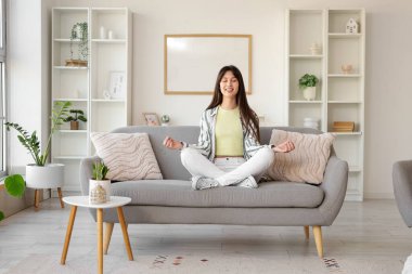 Evdeki kanepede meditasyon yapan genç bir kadın.