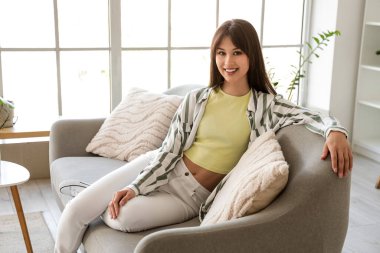 Evdeki gri koltukta oturan gülümseyen genç kadın.