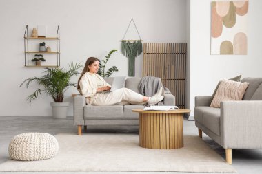 Güzel genç bayan şık oturma odasındaki rahat gri koltukta dizüstü bilgisayar kullanıyor.