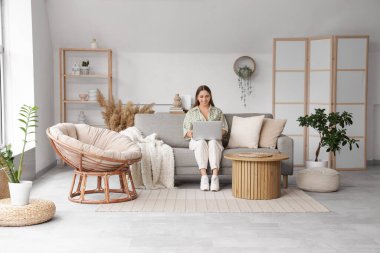 Güzel genç bayan şık oturma odasındaki rahat gri koltukta dizüstü bilgisayar kullanıyor.