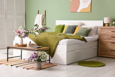 Şık bahar yatak odasının içinde yatak, merdiven, tuvalet ve yeşil duvarın yanındaki masalarda vazolar içinde güzel çiçek buketleri var.