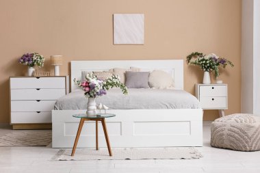 Şık bahar yatak odasının içinde yatak, tuvalet ve güzel çiçek buketleri bej duvarın yanındaki masalarda.