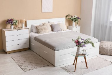 Şık bahar yatak odasının içinde yatak, tuvalet ve güzel çiçek buketleri bej duvarın yanındaki masalarda.