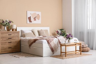 Şık bahar yatak odasının içinde yatağı, aynası, tuvaleti ve bej duvarın yanındaki bankta duran vazolar.