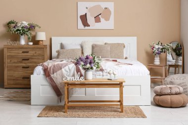 Şık bahar yatak odasının içinde yatağı, aynası, tuvaleti ve bej duvarın yanındaki bankta duran vazolar.