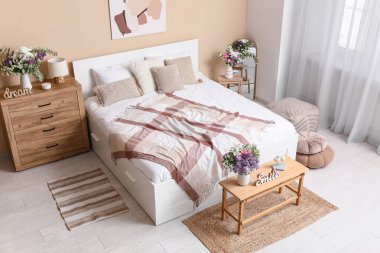 Şık bahar yatak odasının içinde yatak, ayna, tuvalet ve güzel çiçek buketleri bej duvarın yanındaki bankta, üst manzarada.