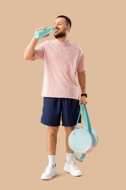 Spor çantalı genç adam bej arka planda su içiyor.
