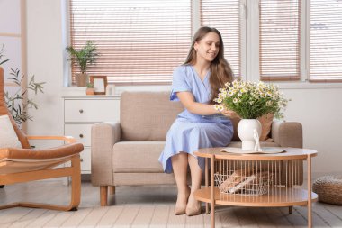 Oturma odasındaki masada papatyaları olan genç bir kadın ve vazo.