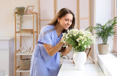 Oturma odasındaki tuvalette papatya çayı ve vazo taşıyan genç bir kadın.