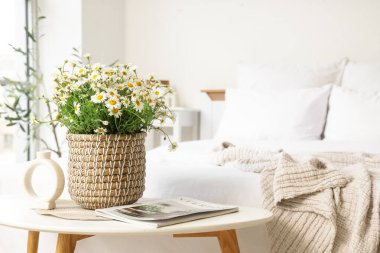 Yatak odasındaki masasında papatya ve dergi olan hasır tencere.