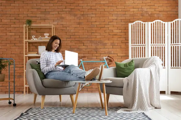 Evdeki yumuşak gri koltukta dizüstü bilgisayar kullanan genç kadın.