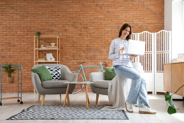 Genç bir kadın evdeki yumuşak gri koltukta dizüstü bilgisayar kullanıyor.