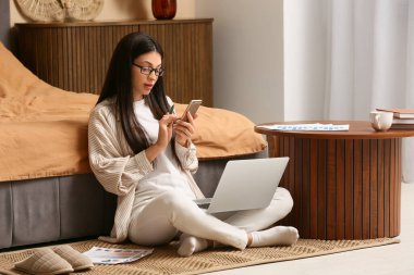 Yatak odasındaki halıda otururken cep telefonu ve dizüstü bilgisayarla çalışan güzel genç bir kadın.