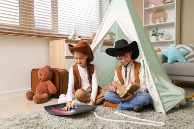 Kovboy gibi giyinmiş şirin çocuklar evdeki çadırda macera kitabı okuyorlar.