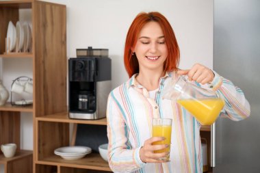 Genç kızıl saçlı kadın mutfakta cama meyve suyu döküyor.