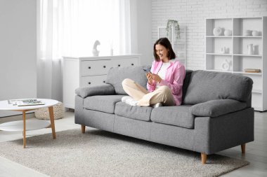 Akıllı ev güvenlik kontrol paneli olan cep telefonunu odasındaki kanepede tutan mutlu genç kadın.