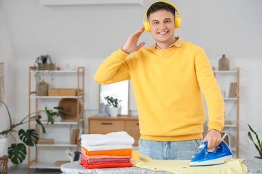 Kulaklıklı yakışıklı genç adam evde giysilerini ütülüyor.