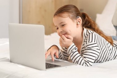 İşitme cihazı olan küçük kız yatak odasında dizüstü bilgisayar kullanıyor.
