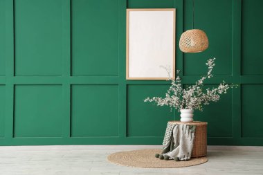 Hasır masada çiçek açan dallar ve oturma odasındaki yeşil duvarın yanında asılı duran çerçeve.