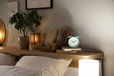 Gece yatak odasının yanındaki rafta parlayan lambalar, kitaplar ve çiçekli çalar saat.