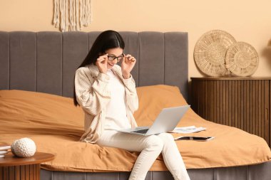 Dizüstü bilgisayarla çalışan ve yatak odasında oturan güzel genç bir kadın.