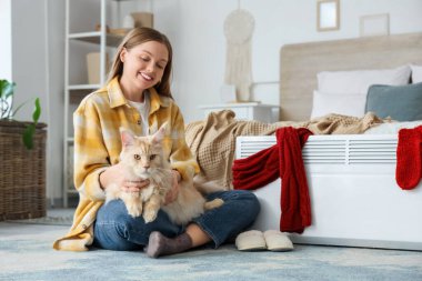 Yatak odasında elektrikli konteyner ısıtıcısının yanında Maine Rakun kedisi olan genç bir kadın oturuyor.