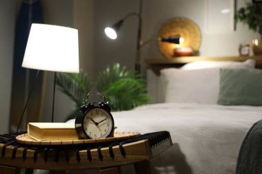 Gece yatak odasının yanındaki hasır masada parlayan lamba ve kitap bulunan çalar saat.
