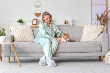Pijamalı, yumuşak terlikli ve evdeki kanepede oturan güzel bir kadın.