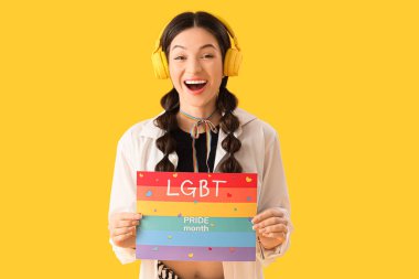 Sarı arka planda LGBT Övünme Ayı yazılı gökkuşağı bayrağı tutan güzel genç bir kadın.