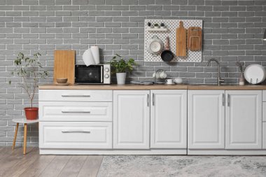 Gri tuğla duvarda beyaz tezgah ve tahta asılı modern mutfağın içi.