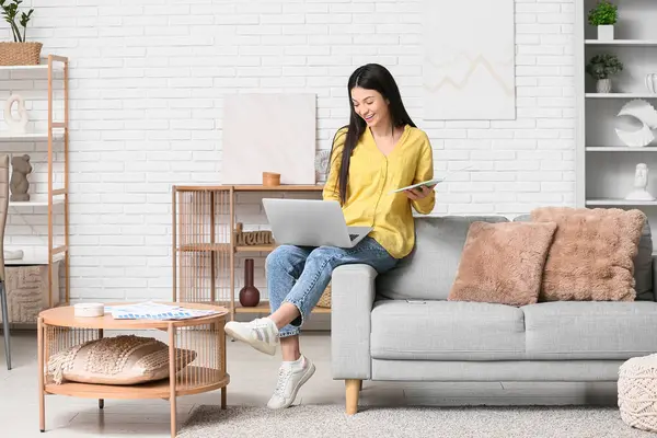 Oturma odasında dizüstü bilgisayar ve defterle çalışan güzel genç bir kadın.