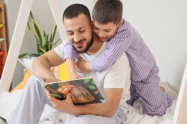 Mutlu baba ve oğlu pijamalarıyla evde kitap okuyorlar.