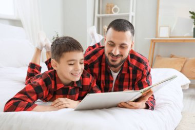 Mutlu baba ve oğlu kareli pijamalarıyla evde yatakta kitap okuyorlar.