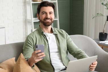 Bilgisayarlı yakışıklı adam evdeki koltukta cep telefonu kullanıyor.