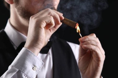 Resmi giyinmiş yakışıklı genç adam siyah arka planda puro yakıyor, yakın plan.
