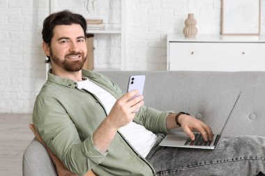 Bilgisayarlı yakışıklı adam evdeki koltukta cep telefonu kullanıyor.