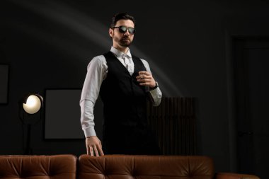 Resmi kıyafetler ve güneş gözlüğü takan yakışıklı genç adam karanlık odada kanepenin yanında puro içiyor.