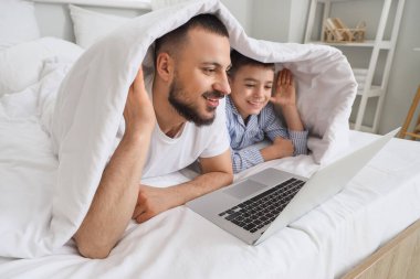 Mutlu baba ve oğlu pijamalarıyla evde dizüstü bilgisayarlarıyla yatıyorlar.