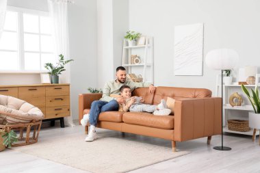 Mutlu baba ve cep telefonlu oğlu evdeki kahverengi deri koltukta oturuyorlar.